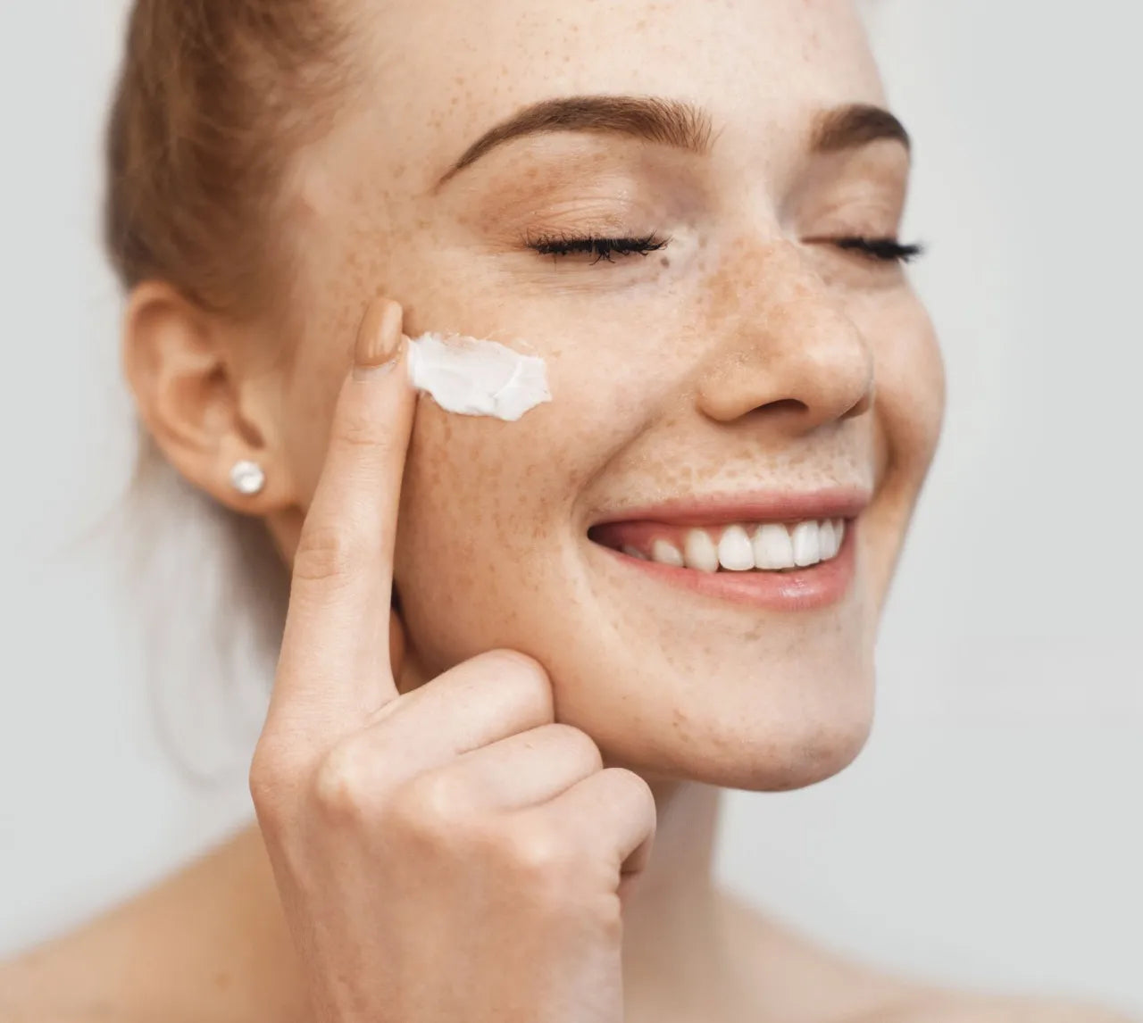 Jeune femme souriante appliquant une crème Derma'Love sur sa joue. Ses taches de rousseur et sa peau naturelle mettent en valeur l'approche douce de nos soins. Son expression joyeuse illustre le plaisir d'utiliser des produits efficaces et respectueux de la peau, promettant un teint éclatant de santé.