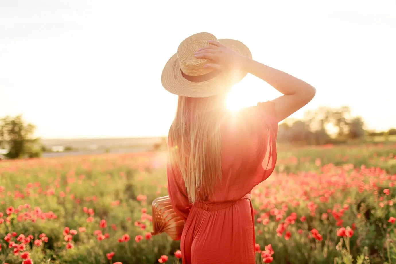 Femme en robe rouge dans un champ de coquelicots au coucher du soleil, tenant son chapeau de paille. Sa silhouette illuminée évoque l'éclat naturel que procurent les soins Derma'Love. L'image symbolise la protection et le rayonnement de la peau, en harmonie avec la nature, grâce à nos produits de beauté.