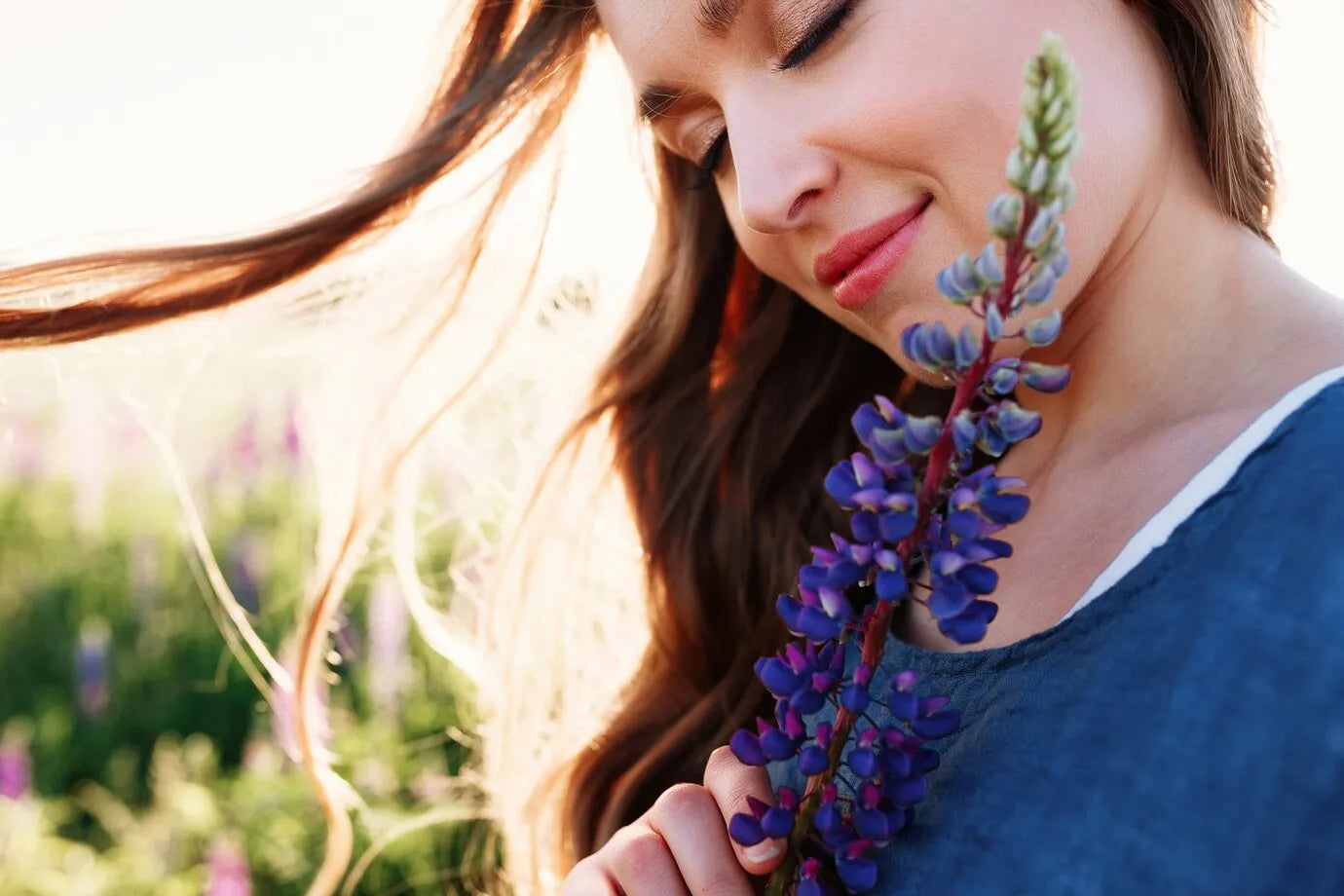 Jeune femme souriante tenant une fleur de lupin, sa peau radieuse illuminée par le soleil couchant. Cette image évoque la beauté naturelle et l'éclat que procurent les soins Derma'Love. L'harmonie entre la fleur et le teint lumineux illustre notre engagement pour des produits respectueux de la peau et de l'environnement.