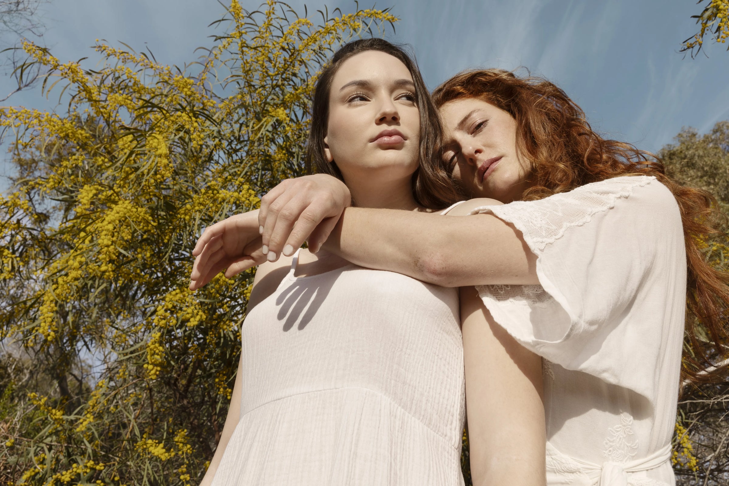 Deux femmes aux peaux éclatantes, vêtues de blanc, s'enlacent devant un mimosa en fleurs. Leurs visages sereins et leur peau lumineuse illustrent les bienfaits des soins Derma'Love pour tous types de peau. L'image évoque la douceur et l'efficacité des produits naturels, en harmonie avec la beauté de la nature environnante.