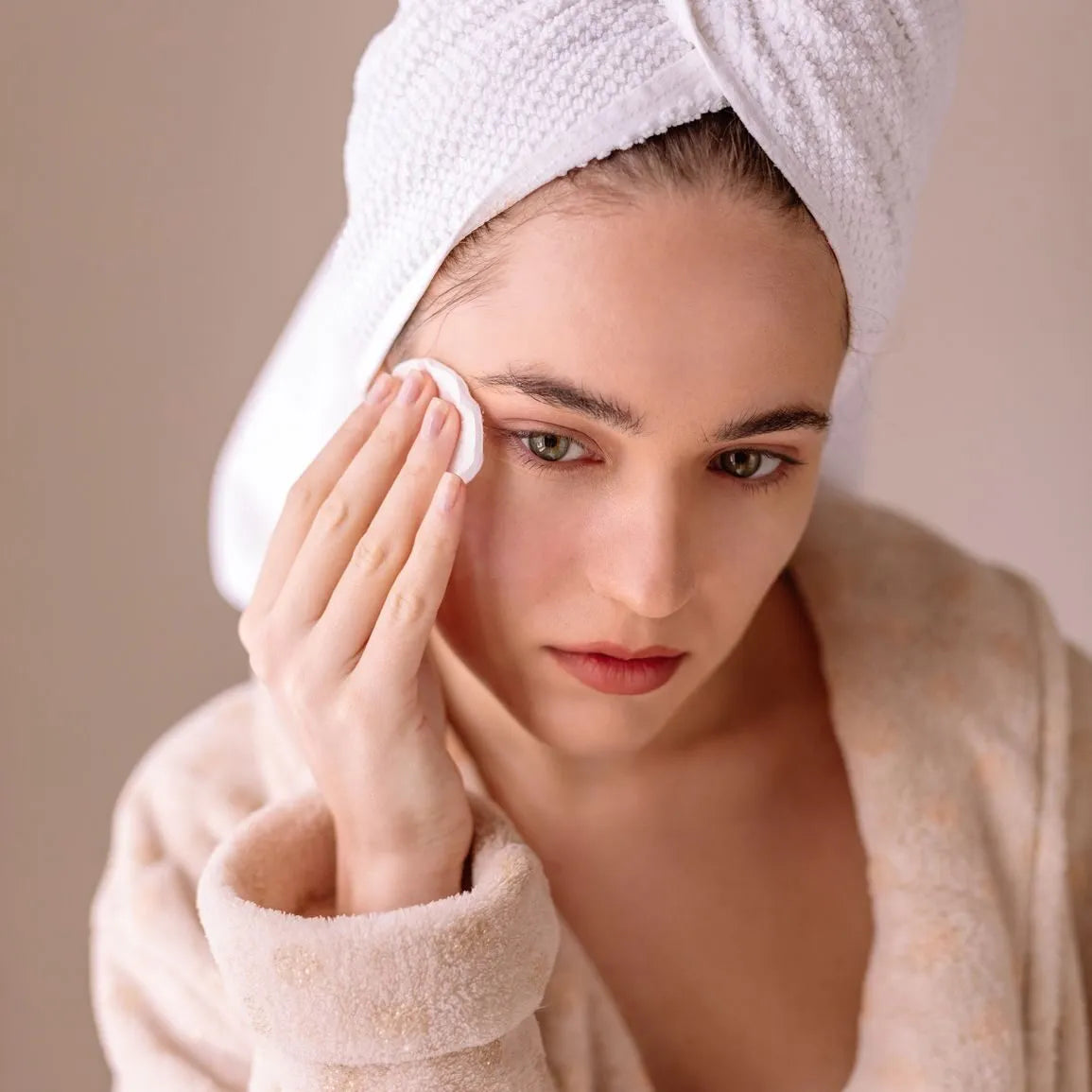 Jeune femme appliquant délicatement un soin Derma'Love sur son visage. Enveloppée dans un peignoir doux et les cheveux enroulés dans une serviette, elle incarne le rituel de beauté quotidien. Son geste précis et son regard concentré illustrent l'importance d'une routine de soins adaptée pour une peau éclatante de santé.